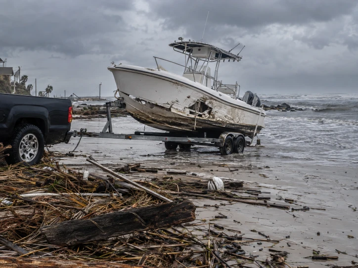 Damaged center-console boat tilted on a trailer beside shoreline debris
