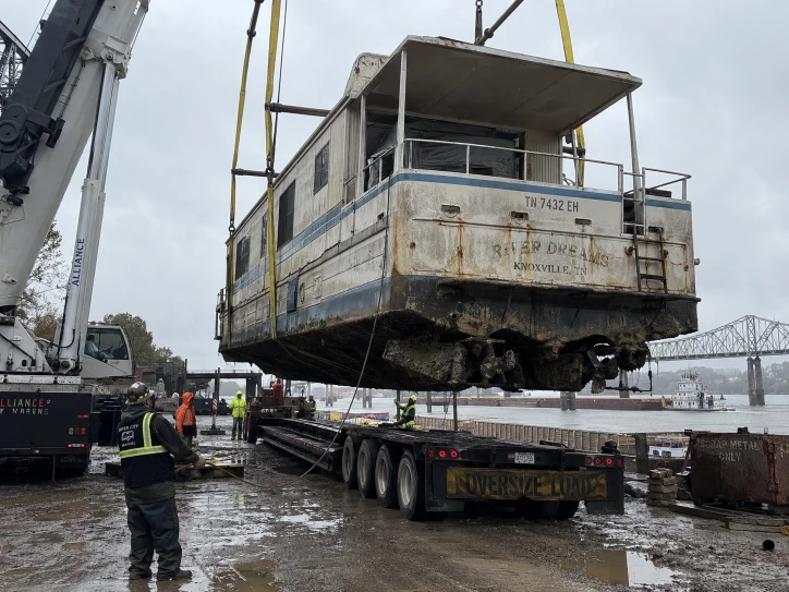 Rear balcony of a weathered houseboat lifted over a long trailer beside a river